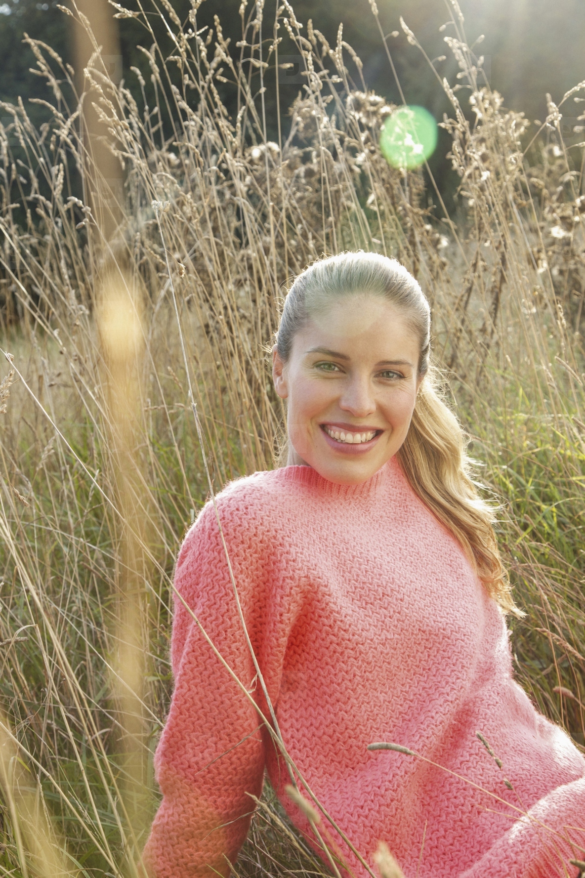 Portrait of beautiful blond woman in pink sweater sitting in sunny tall grass