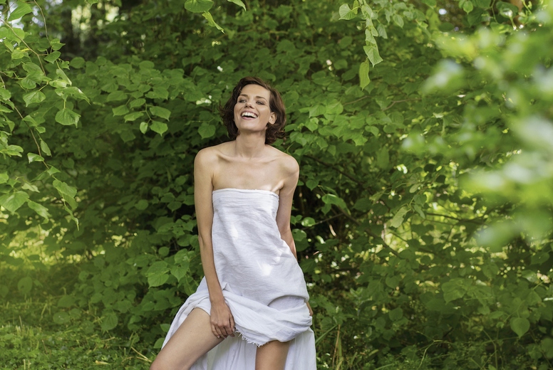 Portrait of tall carefree beautiful woman in strapless dress standing among green leaves in woods