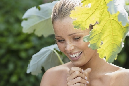 Beautiful young woman with bare shoulders smiling and looking down among green leaves in garden
