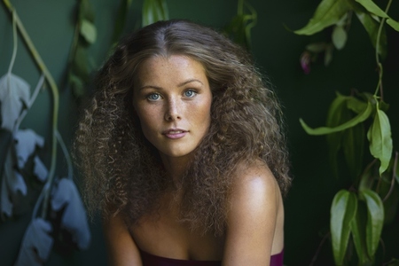 Portrait of beautiful young woman with curly brown hair and blue eyes surrounded by tropical leaves