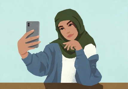 Young Muslim woman in hijab taking selfie with smart phone against blue background