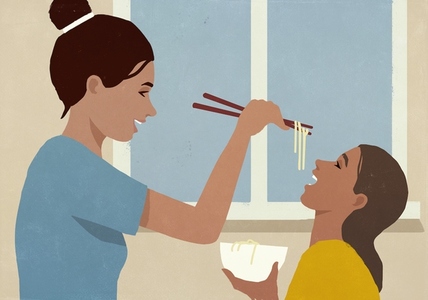 Happy mother feeding noodles to daughter with chopsticks by window at home