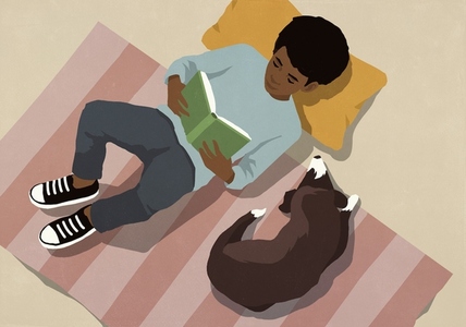 View from above of boy relaxing reading book on rug next to dog at home