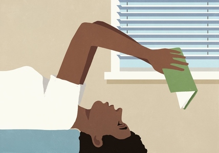 Woman laying over edge of bed and reading book upside down at home