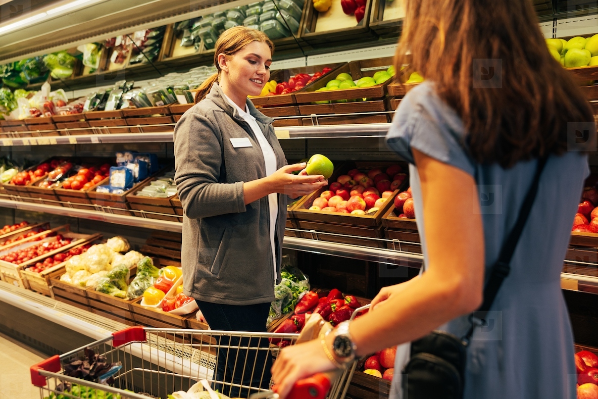 Smiling female worker standing at shelves with fruits and vegetables and showing an apple to a customer