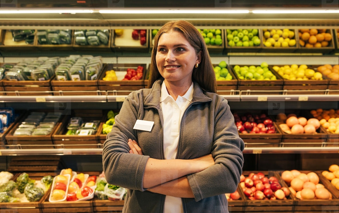 Confident female employee with arms crossed standing against shelves with fruits and vegetables in a grocery store