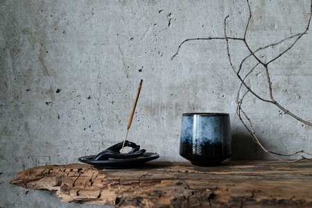 Burning incense stick in a hand shaped holder with a ceramic cup on rustic wood against a concrete wall  Minimalist wabi sabi still life