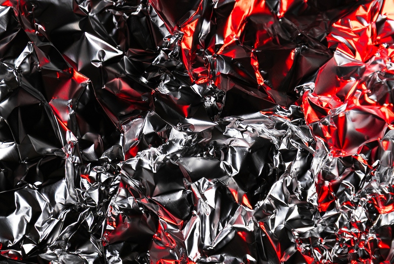 Close up of crumpled aluminum foil with shiny silver surface and dramatic red highlights. Abstract metallic background with reflective texture and artistic light play.