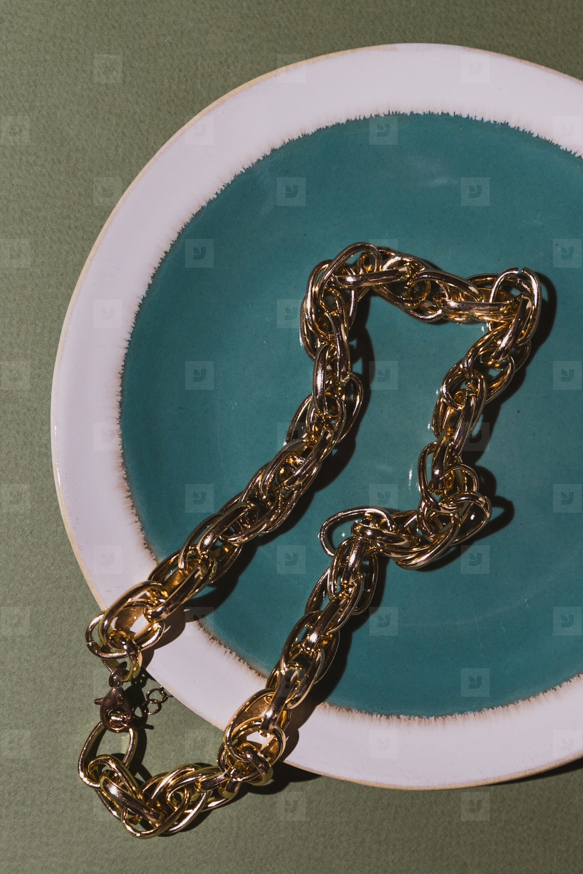 Vintage gold chain displayed in a ceramic plate with teal glaze on a green background  Minimalist still life composition with retro charm