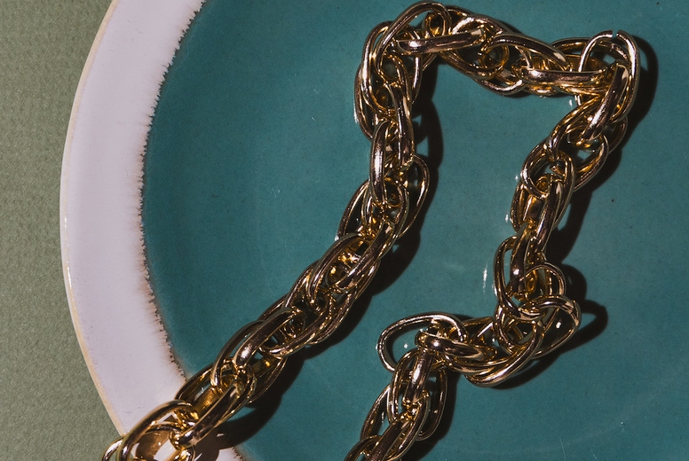 Vintage gold chain displayed in a ceramic plate with teal glaze on a green background. Minimalist still life composition with retro charm