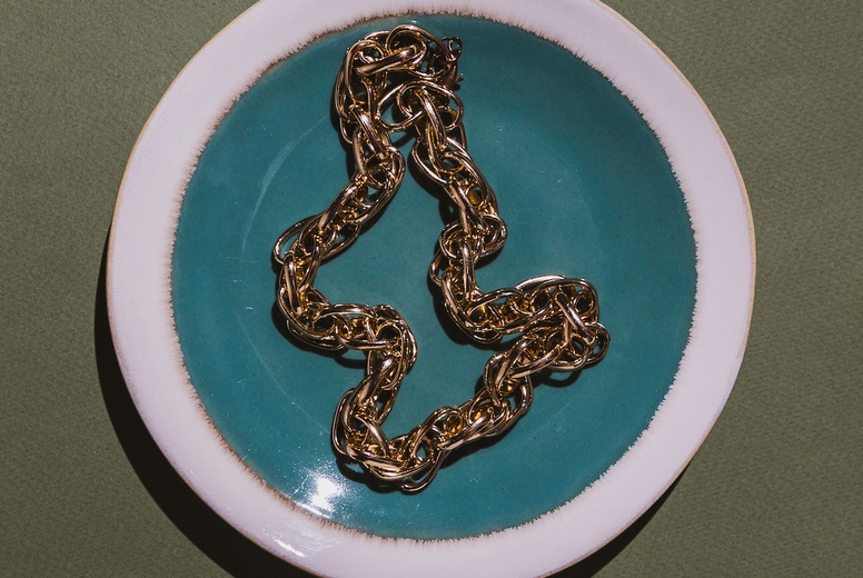 Vintage gold chain displayed in a ceramic plate with teal glaze on a green background. Minimalist still life composition with retro charm