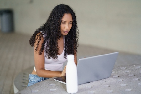 A focused  young woman is diligently working on her laptop with a bottle visible in the background