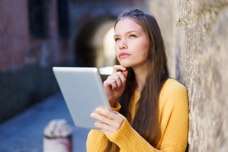A Thoughtful Young Woman Engaged with Her Tablet While Sitting in an Urban Environment A Thoughtful Young Woman Engaged with Her Tablet While Sitting in an Urban Environment
