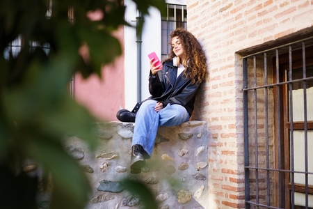 A casually dressed young woman is sitting on a wall in a relaxed posture  using her smartphone