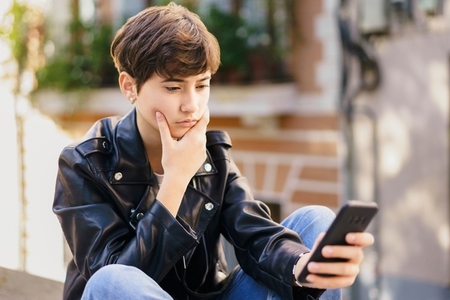 A thoughtful teenage boy is deeply engaged with his smartphone while outdoors in the city