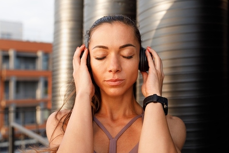 Close up of a young female with closed eyes holding wireless headphones and listening to music after a workout at sunset