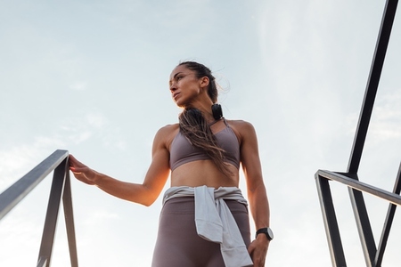 Low angle view of young  slim female in sportswear standing on the stairs on roof and looking away