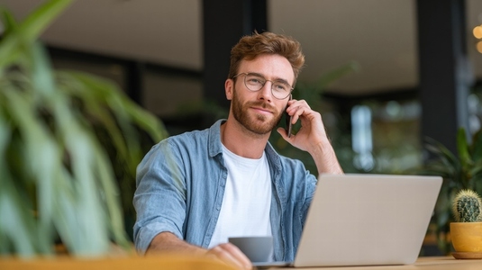 Young entrepreneur working in cafe with laptop  talking on phone  relaxed  natural light
