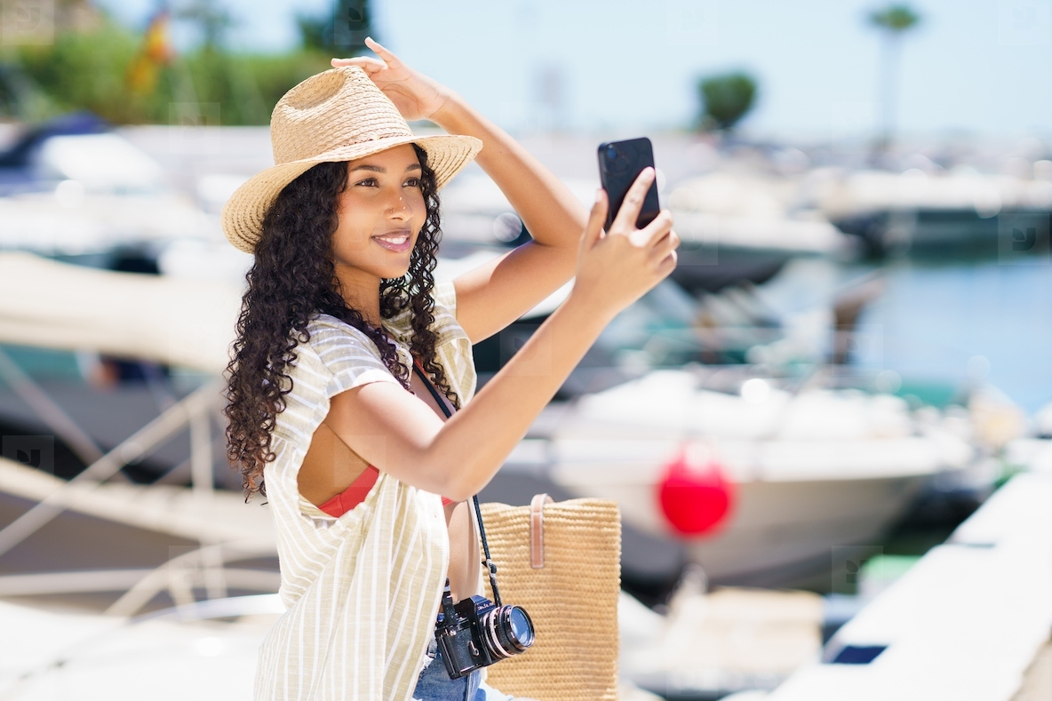 A cheerful young woman takes a selfie at a vibrant marina on a bright sunny day