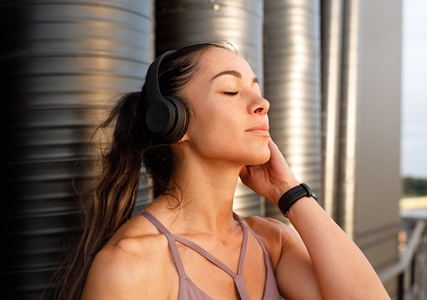 Young female with closed eyes wearing wireless headphones standing at sunset outdoors and enjoying a break during a workout