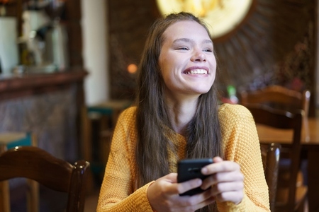 A cheerful young woman is smiling widely while she is using her smartphone in a cozy cafe A cheerful young woman is smiling widely while she is using her smartphone in a cozy cafe