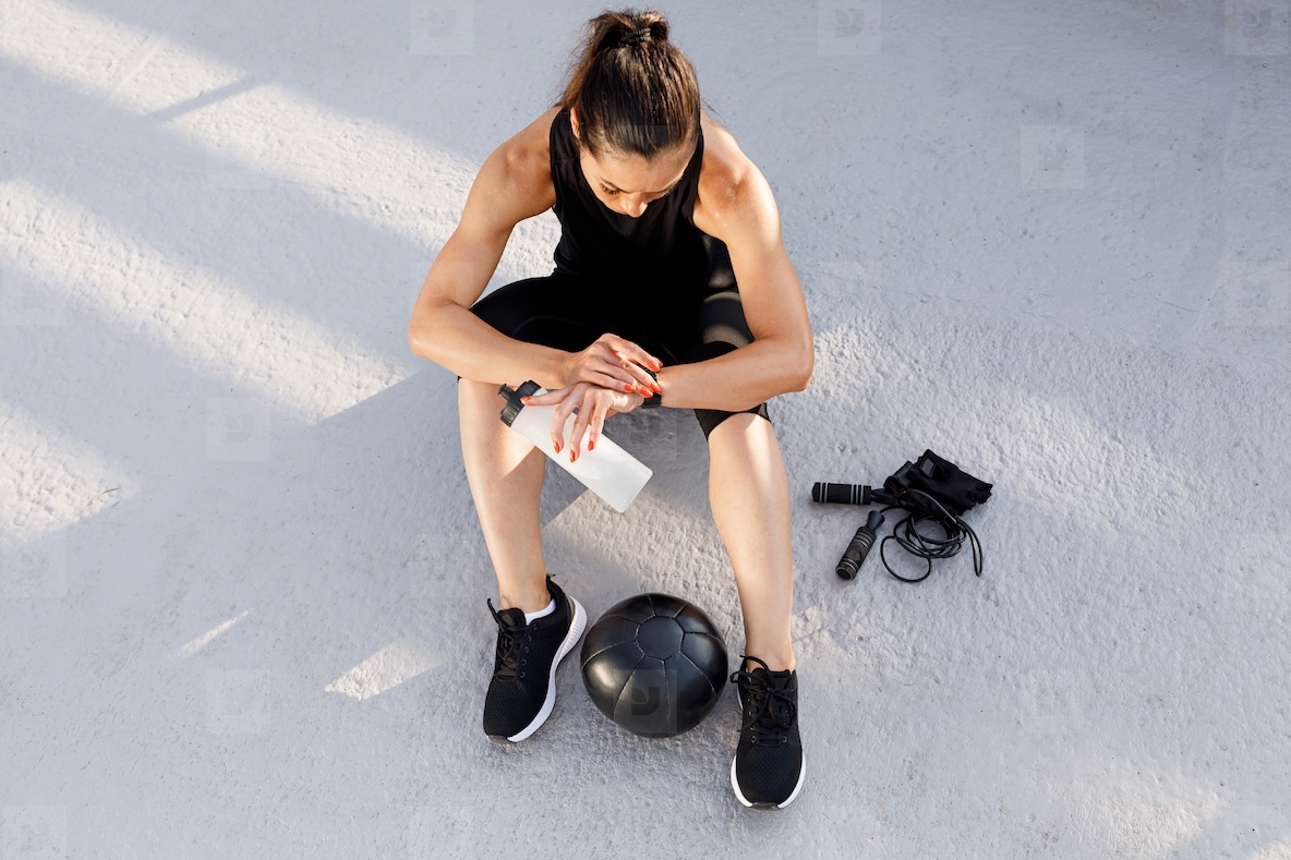High angle of a young slim sportswoman holding a water bottle and looking at her smartwatch while sitting