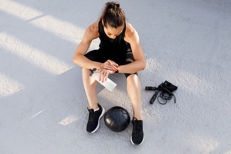 High angle of a young slim sportswoman holding a water bottle and looking at her smartwatch while sitting