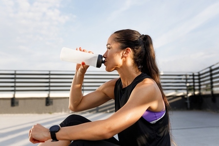 Side view of a young female in sportswear drinking water from a white bottle while sitting on the roof