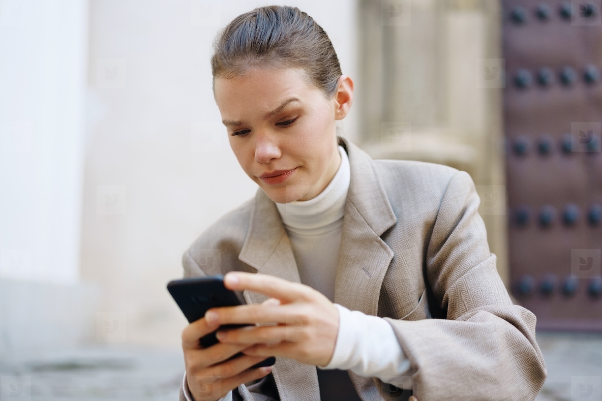 A young woman is sitting outside in an urban setting engaged with her smartphone device