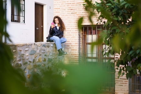 A Young Woman with Beautiful Curly Hair is Sitting on a Wall in an Urban Environment