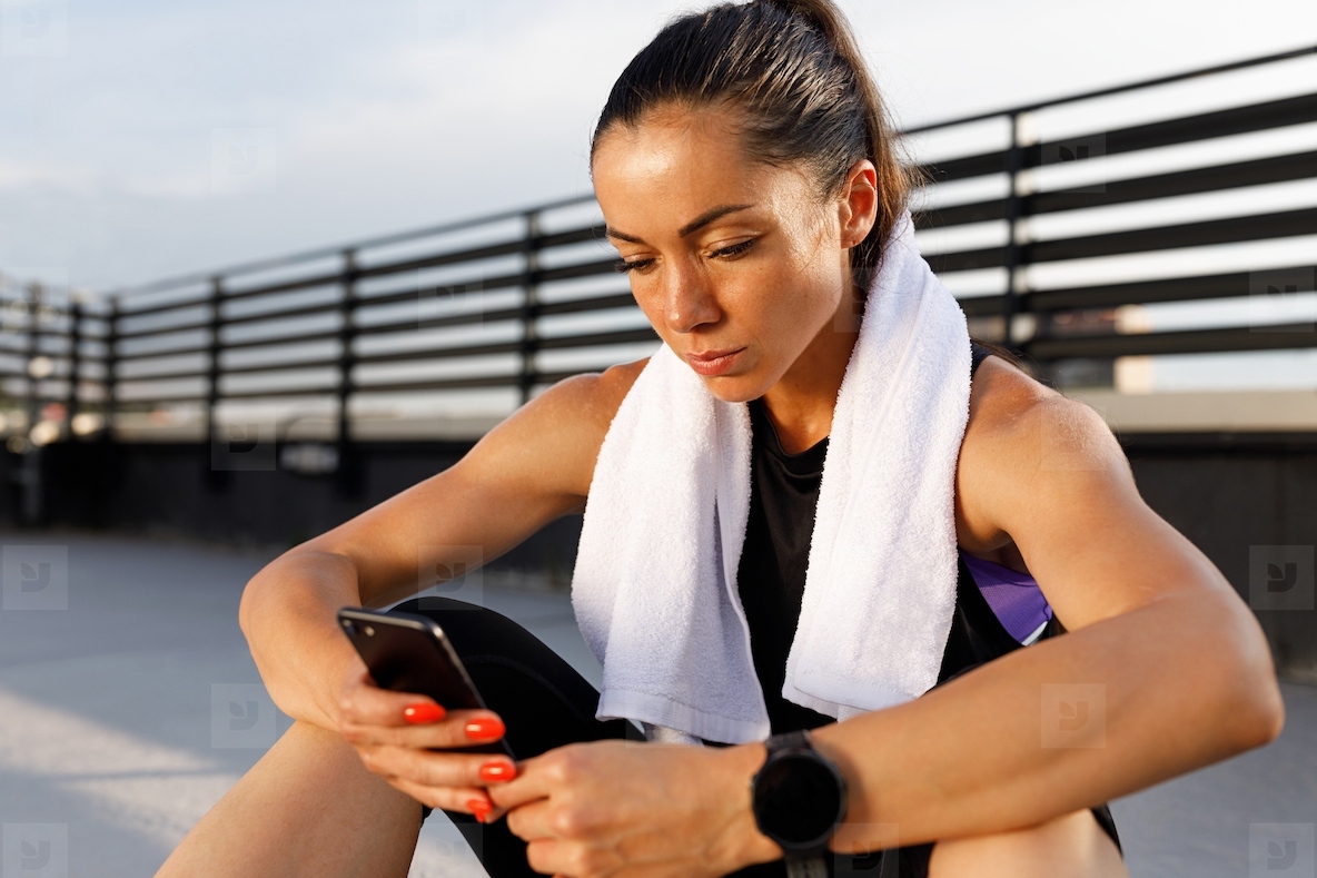 Slim athlete with a white towel on her neck using a phone while taking a break during a workout