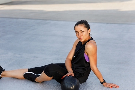 Young sportswoman relaxing on the roof during workout  Female in sportswear sitting on the roof with medicine ball