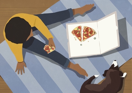 View from above boy eating pizza from box on floor next to dog