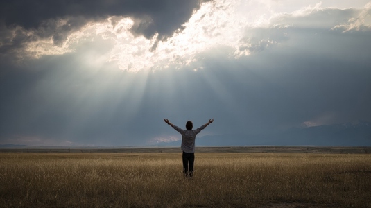 Person stands in wide open field  arms raised towards sky  expressing joy and freedom under dramatic clouds and sunlight