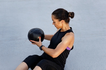 Muscular young female with medicine ball practicing outdoors  Woman in black fitness attire exercising using medicine ball while sitting