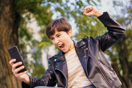 A very excited young person joyfully celebrating while using their mobile phone outdoors in a park