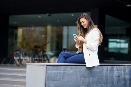 A young woman is sitting outdoors  happily texting on her phone while enjoying the day