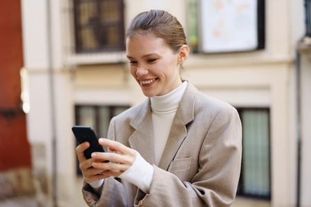 A Smiling Woman Using Her Smartphone Outdoors While Wearing a Stylish and Fashionable Outfit