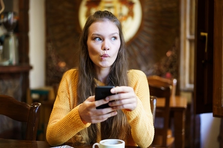 A Thoughtful and Young Woman Happily Enjoying a Delicious Coffee While Using Her Smartphone