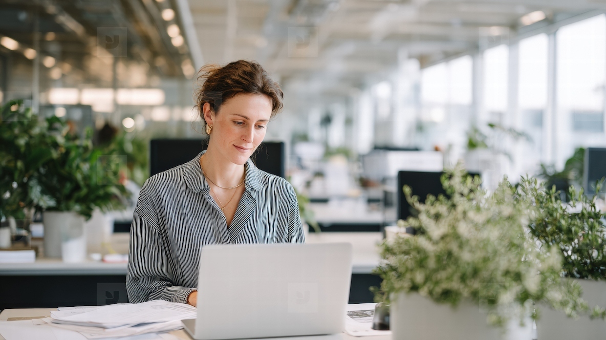 Focused employee working modern office desk with laptop  surrounded by plants and natural light  creating productive