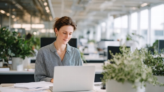 Focused employee working modern office desk with laptop  surrounded by plants and natural light  creating productive