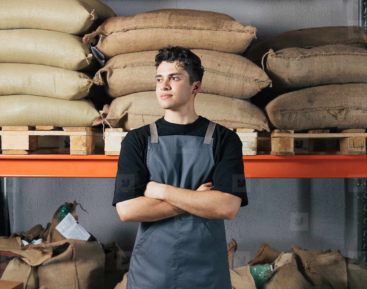 Confident barista standing at warehouse with arms crossed  Male worker in apron standing against shelf with bags at warehouse