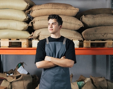 Confident barista standing at warehouse with arms crossed  Male worker in apron standing against shelf with bags at warehouse