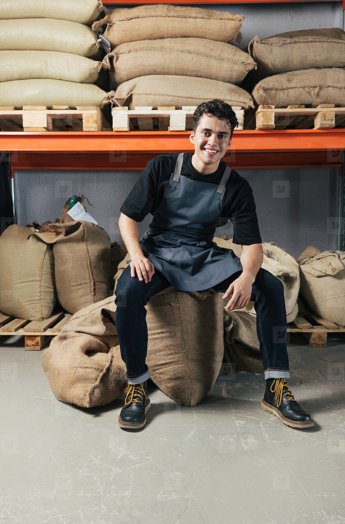 Exhausted warehouse worker sitting on a coffee bag and looking at camera  Young worker in apron sitting in warehouse