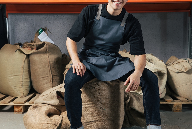 Exhausted warehouse worker sitting on a coffee bag and looking at camera. Young worker in apron sitting in warehouse.