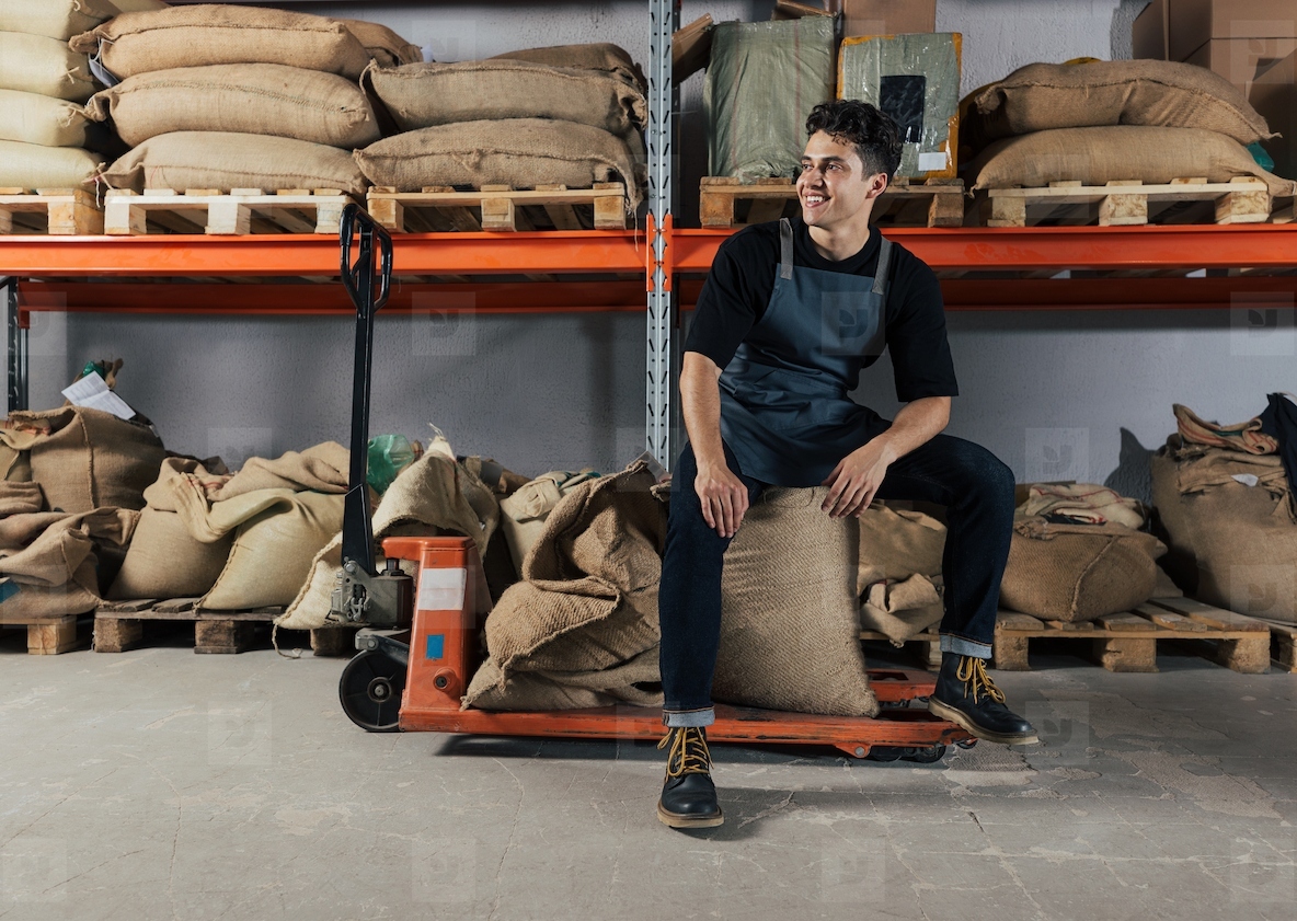 Young smiling warehouse worker sitting on bags and a manual pallet truck Barista at the warehouse relaxing during manual work
