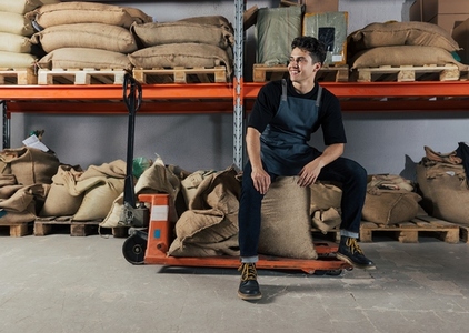 Young smiling warehouse worker sitting on bags and a manual pallet truck  Barista at the warehouse relaxing during manual work