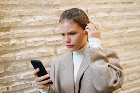 A young woman stands by a textured wall  using her smartphone and showcasing her style