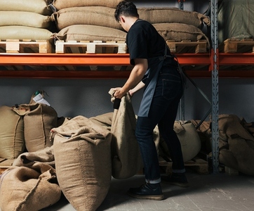 Back view of a young warehouse worker moving a bag Barista is managing bags of coffee next to the shelves Back view of a young warehouse worker moving a bag Barista is managing bags of coffee next to the shelves