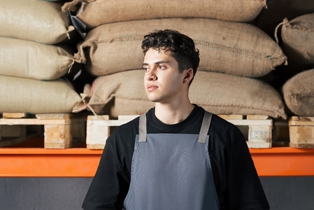 Portrait of a young male wearing an apron standing in a warehouse against bags of coffee beans Portrait of a young male wearing an apron standing in a warehouse against bags of coffee beans